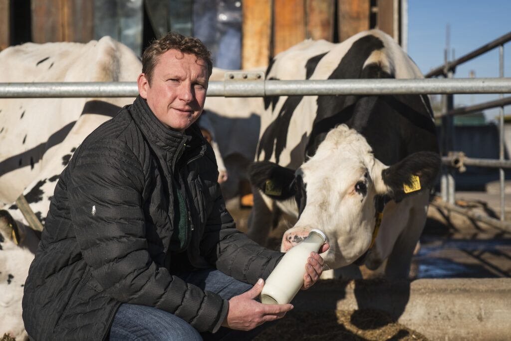 Das Foto zeigt Bauer Thomas mit einer Glasflasche Milch in der Hand. Hinter ihm steht eine Kuh.